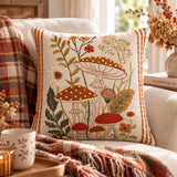 Cuscino Marigold panna con funghi e motivi botanici ricamati, su un divano con plaid autunnale.
