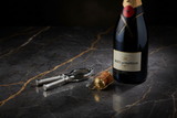 Pinza apri tappo spumante su marmo scuro con bottiglia di champagne e muselet dorato